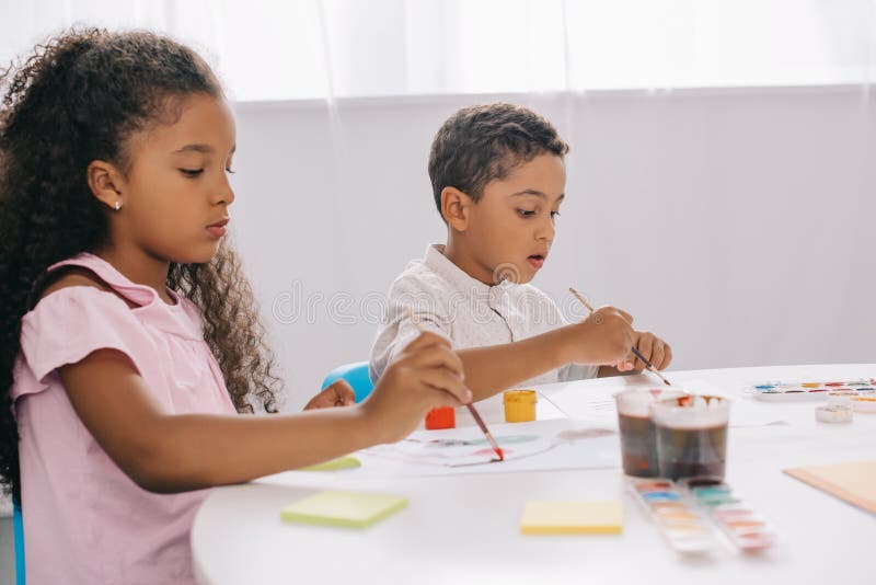 focused african american kids drawing pictures with paints and paint brushes in classroom