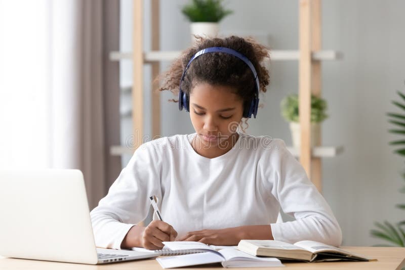 Focused african african teen girl wearing headphones writing notes studying