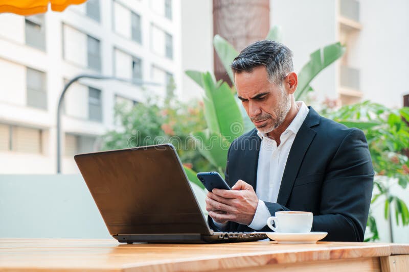 Focused businessman in a suit using a laptop and smartphone while working remotely at a terrace cafe royalty free stock photo