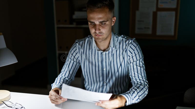 A focused young man reviews documents at night in a modern office setting, exemplifying professionalism and dedication stock image