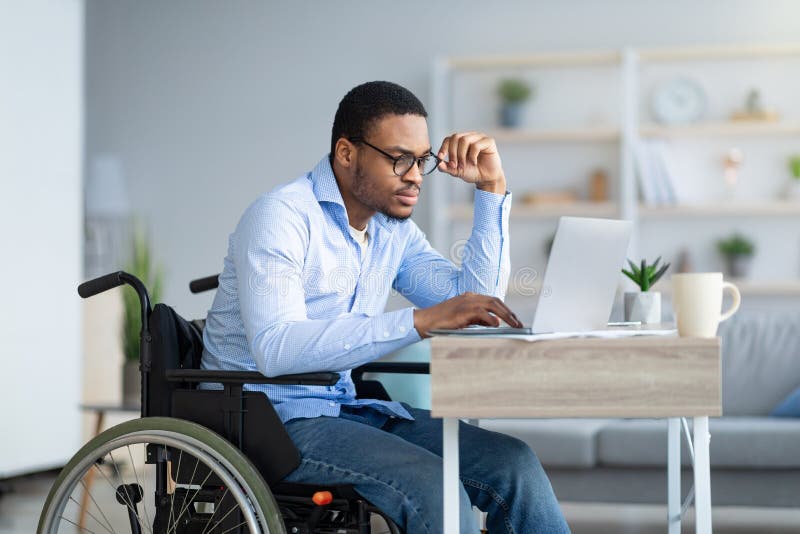 Focused young man in wheelchair using laptop computer for online work or communication at home. Hardworking disabled black guy having webinar or remote