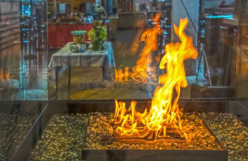 Fogo dançante numa caixa de fogo de vidro com mesas e cadeiras irreconhecíveis, embaçadas e escuras no fundo refletidas no vidro imagens de stock royalty free Fogo dançante numa caixa de fogo de vidro com mesas e cadeiras irreconhecíveis, embaçadas e escuras no fundo refletidas no vidro imagens de stock royalty free