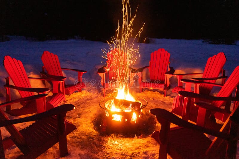 Cozy/romantic evening scene of warming bonfire on snow in winter with red chairs around. Canadian symbols of bear, paw and tree on the fire container. Banff National Park, Alberta, Canada. Cozy/romantic evening scene of warming bonfire on snow in winter with red chairs around. Canadian symbols of bear, paw and tree on the fire container. Banff National Park, Alberta, Canada