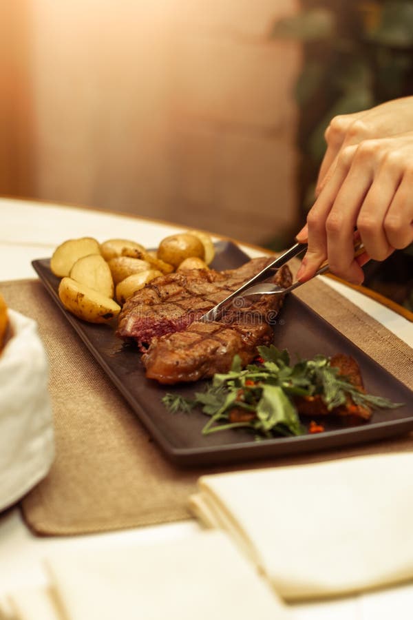 Fokus auf dem Fleisch wo die Gabel und das Messer Schöne weibliche Hände in einem Restaurant am Tabellenschnitt-Rindfleisch strip lizenzfreies stockbild Fokus auf dem Fleisch wo die Gabel und das Messer Schöne weibliche Hände in einem Restaurant am Tabellenschnitt-Rindfleisch strip lizenzfreies stockbild