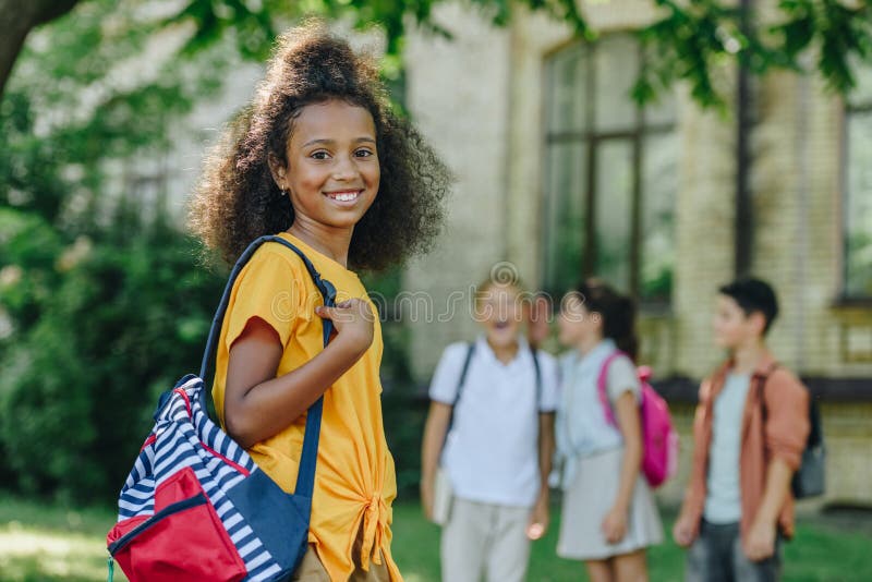 Fokus på den heliga afrikanska amerikanska skolflickan med ryggsäck som leker vid kamera nära multietniska vänner arkivbild