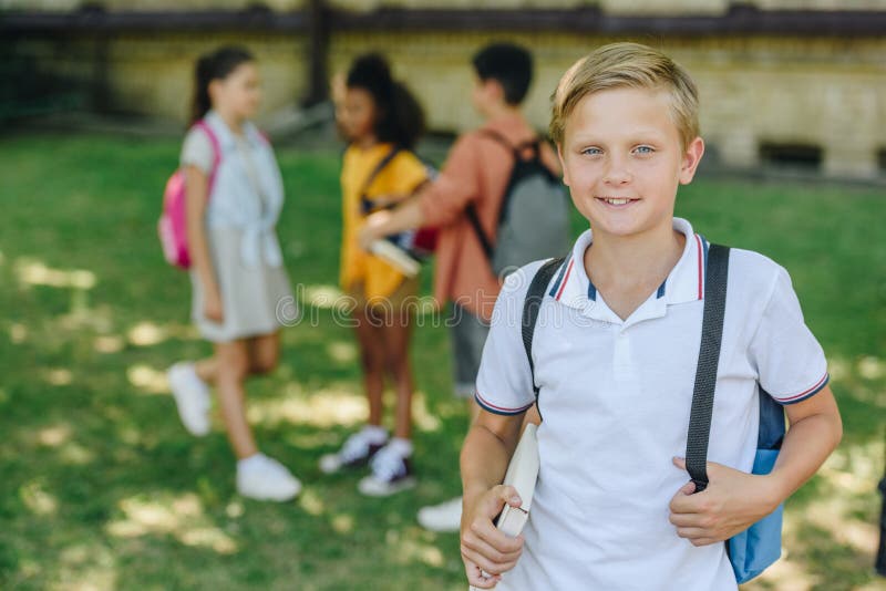 Fokus på leende skolpojke som tittar på kameran medan man står nära mångkulturella vänner arkivbild