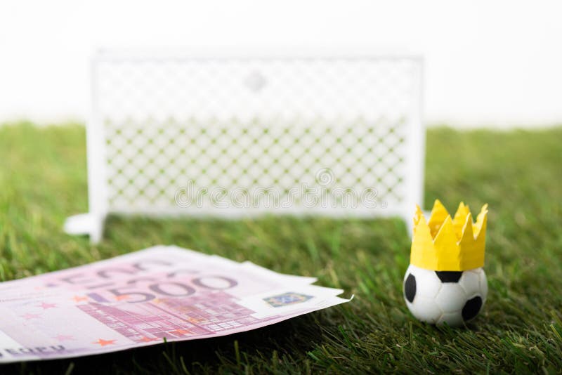Selective focus of toy soccer ball with paper crown near euro banknotes and miniature gates isolated on white, sports betting concept. Selective focus of toy soccer ball with paper crown near euro banknotes and miniature gates isolated on white, sports betting concept