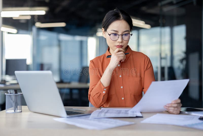 A businesswoman in a modern office setting closely examines paperwork. She is thoughtful, displaying focus and professionalism with technology around. A businesswoman in a modern office setting closely examines paperwork. She is thoughtful, displaying focus and professionalism with technology around.