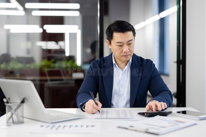 A professional businessman dressed in a blue suit is focused on reviewing financial documents at a modern workspace, surrounded by graphs and a laptop. A professional businessman dressed in a blue suit is focused on reviewing financial documents at a modern workspace, surrounded by graphs and a laptop.