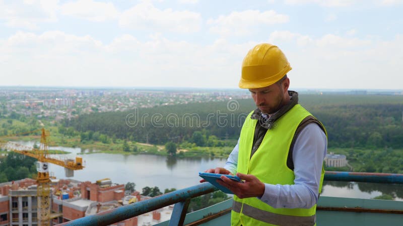 Fokuserad kaukasisk ung man i uniform och hjälm som arbetar med digital tablett medan han står på byggstället arkivfoto