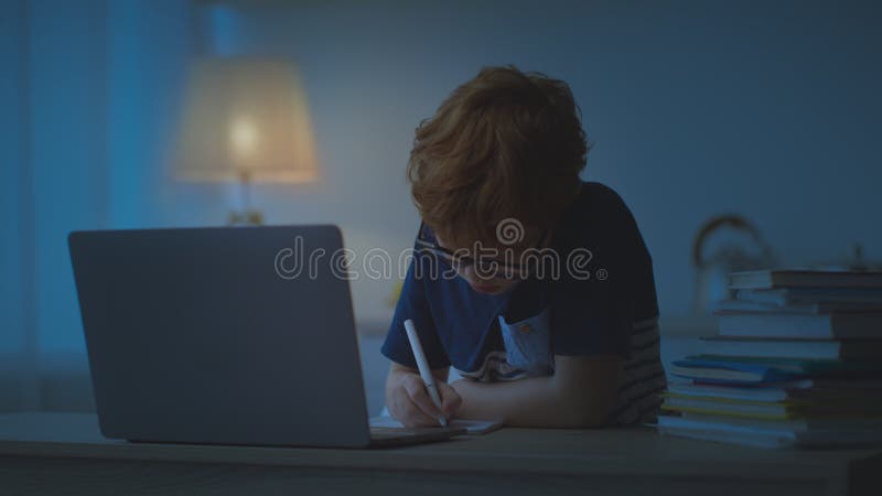 Fokussierter kleiner Schuljunge in Brille, der nachts Hausaufgaben macht, gegen Laptop sitzt, in einem dunklen Raum sitzt und schr stock video footage