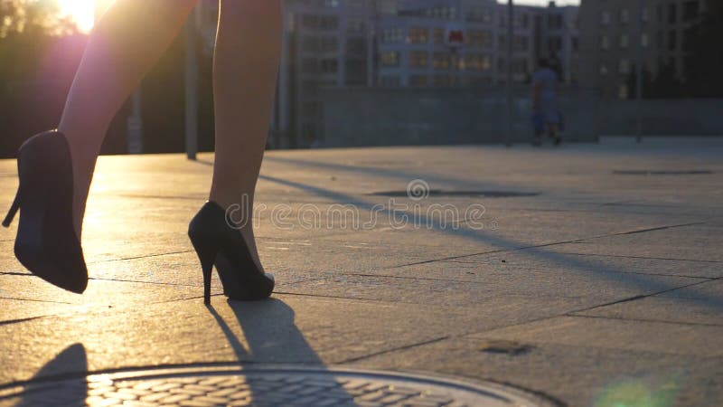 Follow to female legs in high heels shoes walking along city street in sunset time. Feet of business woman in high