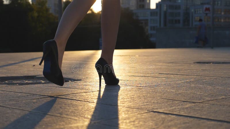 Follow to female legs in high heels shoes walking along city street in sunset time. Feet of business woman in high