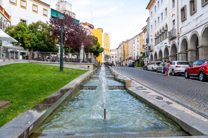 Fontana con acqua a cascata lungo la strada pavimentata con panchine di alberi ed edifici bianchi giallastre immagini stock libere da diritti