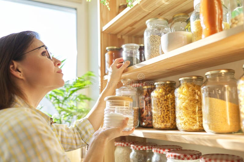 Food storage in pantry, woman holding jar of sugar in hand. Pantry interior, wooden shelf with food cans and kitchen utensils