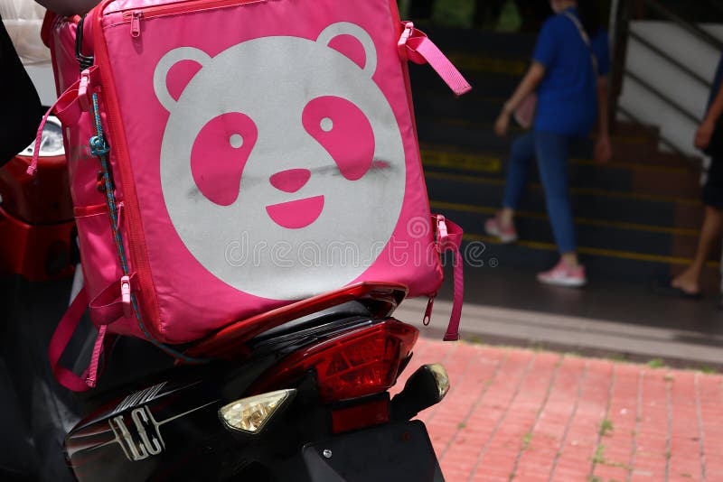 Foodpanda logo on a delivery rider`s squarish container bag at the back of a motorbike in Malaysia. In the background are some pedestrians walking along the pavement of a building. Foodpanda logo on a delivery rider`s squarish container bag at the back of a motorbike in Malaysia. In the background are some pedestrians walking along the pavement of a building