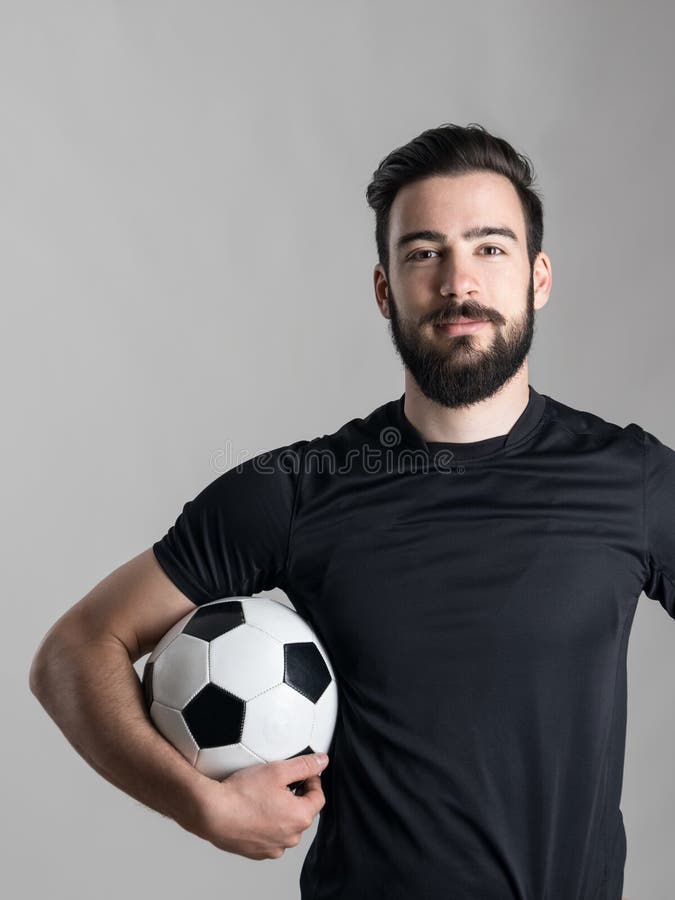 Friendly smiling bearded soccer player holding ball under his arm looking at camera over gray studio background. Friendly smiling bearded soccer player holding ball under his arm looking at camera over gray studio background.