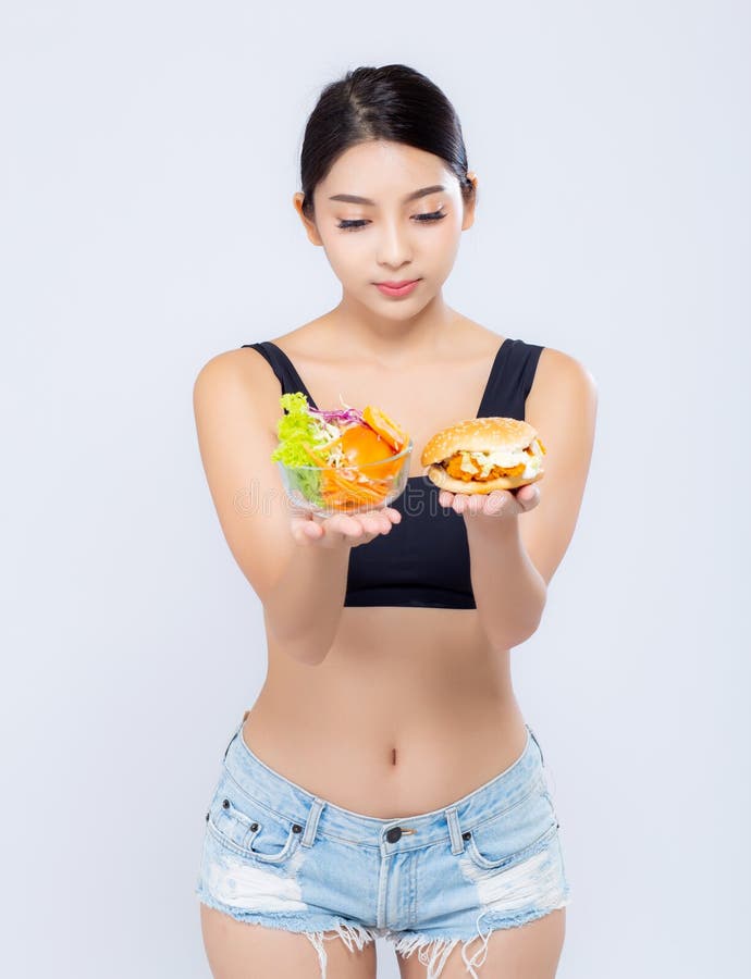 Forma magro da mulher asiática nova bonita com a dieta que escolhe o vegetal de salada fresco e o Hamburger isolados no fundo bra imagem de stock royalty free Forma magro da mulher asiática nova bonita com a dieta que escolhe o vegetal de salada fresco e o Hamburger isolados no fundo bra imagem de stock royalty free