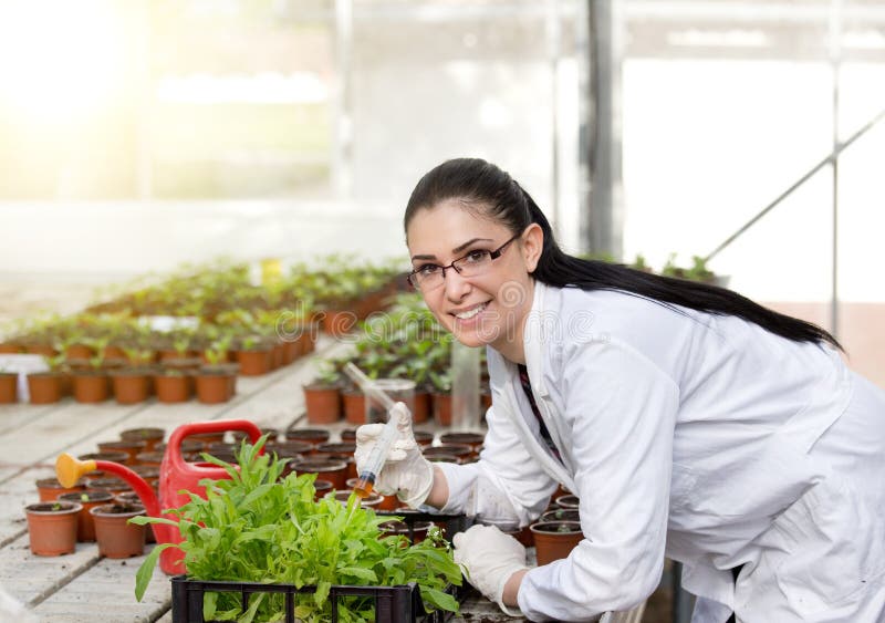 Young woman in white coat testing sprout growth in flower pots in greenhouse. Looking at camera and smiling. Young woman in white coat testing sprout growth in flower pots in greenhouse. Looking at camera and smiling