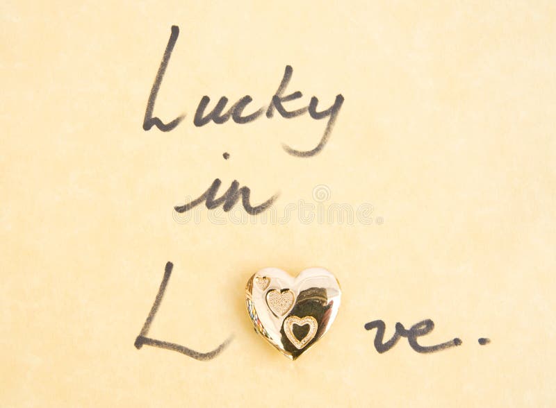 The words ' lucky in love ' with a silver locket replacing the o in love. The image is on straw colored parchment paper. The words ' lucky in love ' with a silver locket replacing the o in love. The image is on straw colored parchment paper.