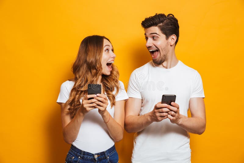 Photo of positive excited people men and women screaming and looking at each other while both using mobile phones isolated over yellow background. Photo of positive excited people men and women screaming and looking at each other while both using mobile phones isolated over yellow background