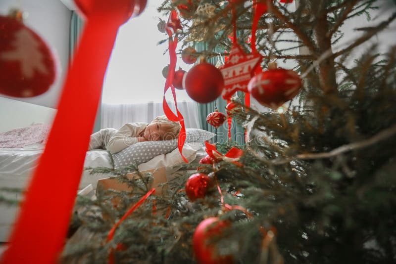Foto av en pojke i pyjamas på en säng bredvid ett julgransträd som dekorerats med röda leksaker arkivfoto