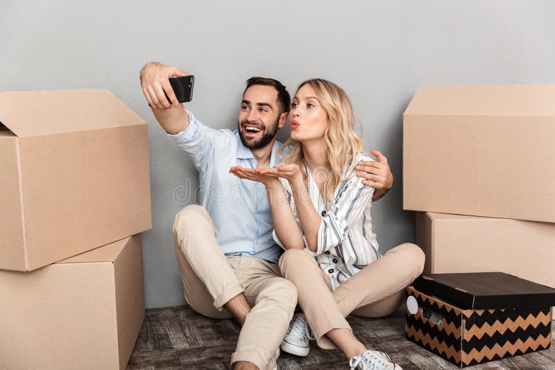 Photo of attractive couple seating near cardboard boxes and taking selfie photo on cellphone isolated over gray wall. Photo of attractive couple seating near cardboard boxes and taking selfie photo on cellphone isolated over gray wall