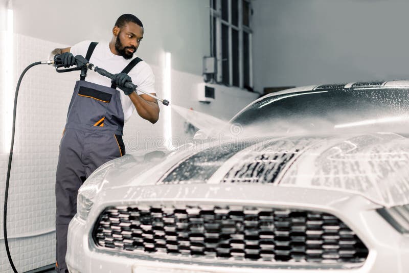 Foto de jovem trabalhador de pele escura em camiseta branca e macacão cinza, lavando carro esportivo branco sob alta pressão imagens de stock royalty free Foto de jovem trabalhador de pele escura em camiseta branca e macacão cinza, lavando carro esportivo branco sob alta pressão imagens de stock royalty free