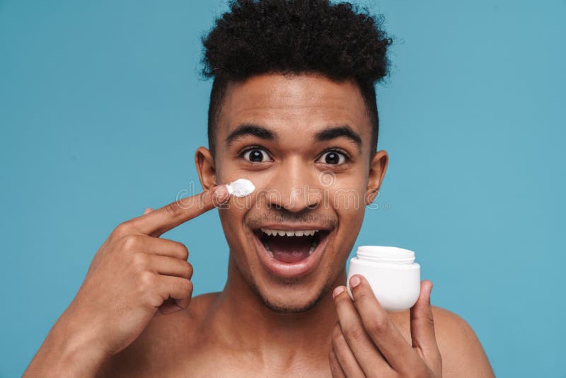 Foto de un hombre afroamericano sin camisa sonriendo y aplicando crema facial imagenes de archivo