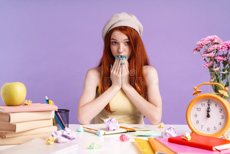 Foto de una estudiante pelirroja cubriéndose la boca mientras estaba sentada en el escritorio fotos de archivo