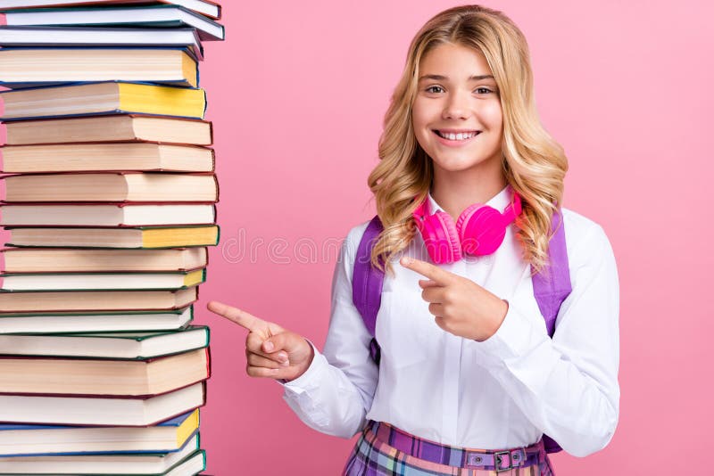 Photo of cute sweet schoolgirl wear formal outfit rucksack headphones pointing book stack smiling isolated pink color background. Photo of cute sweet schoolgirl wear formal outfit rucksack headphones pointing book stack smiling isolated pink color background.