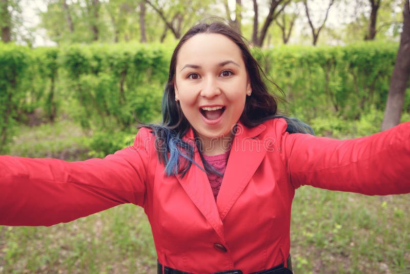 Foto del primo piano Una giovane donna in vestiti rossi sulla via prende un selfie, esamina la macchina fotografica ed i sorrisi fotografia stock libera da diritti Foto del primo piano Una giovane donna in vestiti rossi sulla via prende un selfie, esamina la macchina fotografica ed i sorrisi fotografia stock libera da diritti