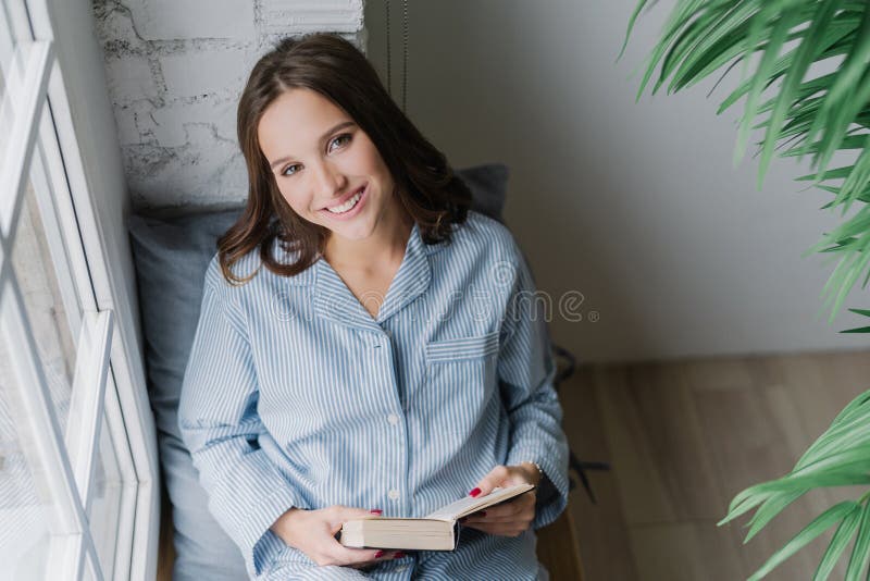 Foto der netten Frau liest romantische Geschichte, hält Buch, trägt zufällige Kleidung, aufwirft auf Fensterbrett im gemütlichen lizenzfreies stockfoto Foto der netten Frau liest romantische Geschichte, hält Buch, trägt zufällige Kleidung, aufwirft auf Fensterbrett im gemütlichen lizenzfreies stockfoto