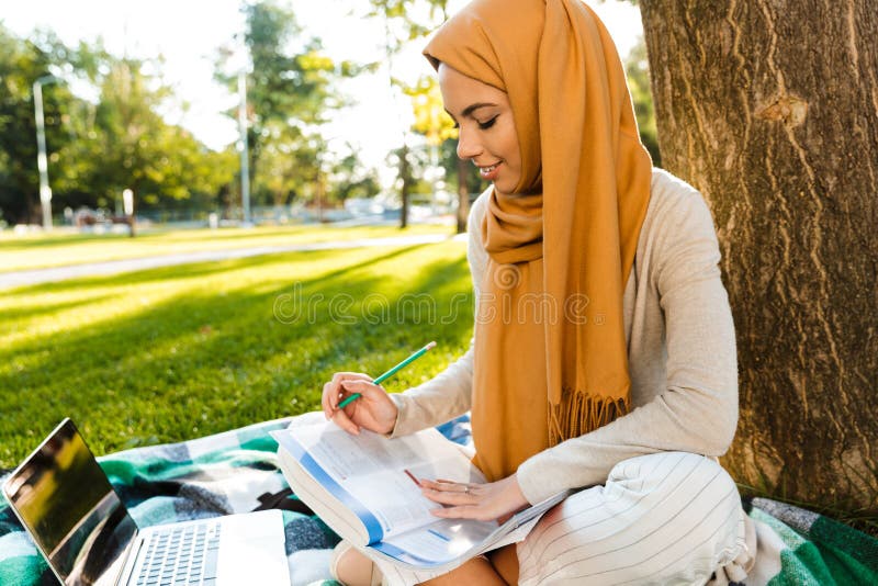 Foto des tragenden Kopftuches des brunette arabischen Studenten, das auf Decke im grünen Park sitzt