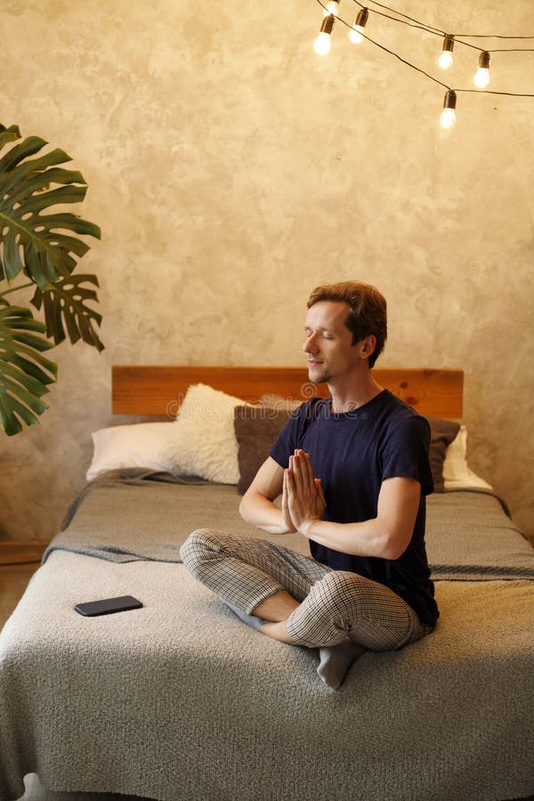 Foto di un americano calmo che medita con un gesto di nome mentre sta seduto sul letto in una stanza buia immagine stock