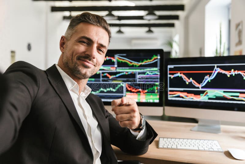 Photo of handsome man 30s wearing suit taking selfie while working in office on computer with graphics and charts at screen. Photo of handsome man 30s wearing suit taking selfie while working in office on computer with graphics and charts at screen