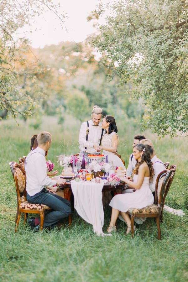 A foto dos recém-casados que cortam sua primeira parte de bolo de casamento junto, quando os convidados a olharem E foto de stock royalty free A foto dos recém-casados que cortam sua primeira parte de bolo de casamento junto, quando os convidados a olharem E foto de stock royalty free
