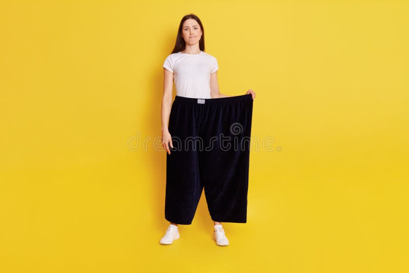 Full length photo of Young adult Caucasian girl wearing casual t shirt and too big size pants, looks at camera with puzzled expression and bewilderment, purses lips, isolated over yellow background. Full length photo of Young adult Caucasian girl wearing casual t shirt and too big size pants, looks at camera with puzzled expression and bewilderment, purses lips, isolated over yellow background