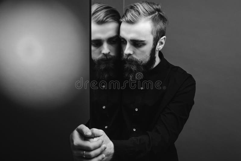 Foto preto e branco de um homem com uma barba e um penteado à moda vestidos na posição preta da camisa ao lado do espelho fotos de stock Foto preto e branco de um homem com uma barba e um penteado à moda vestidos na posição preta da camisa ao lado do espelho fotos de stock
