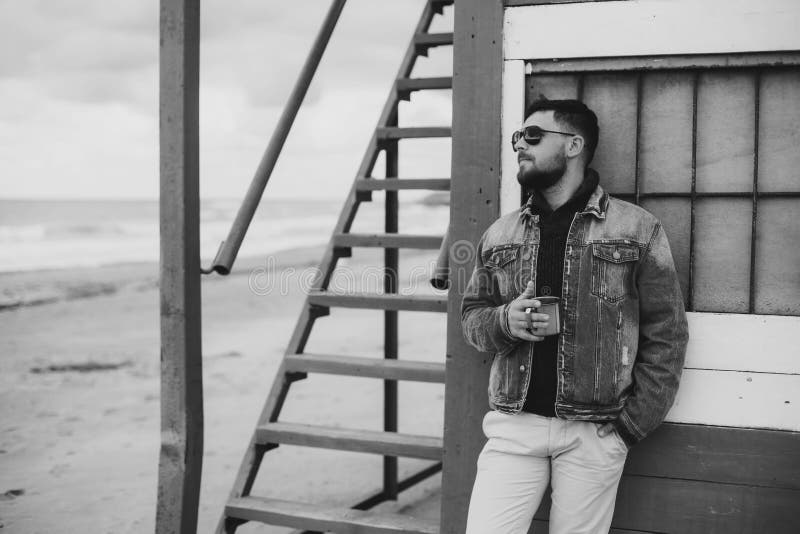 Foto preto e branco de um homem novo à moda com uma barba que está o oceano próximo com um copo do chá foto de stock Foto preto e branco de um homem novo à moda com uma barba que está o oceano próximo com um copo do chá foto de stock