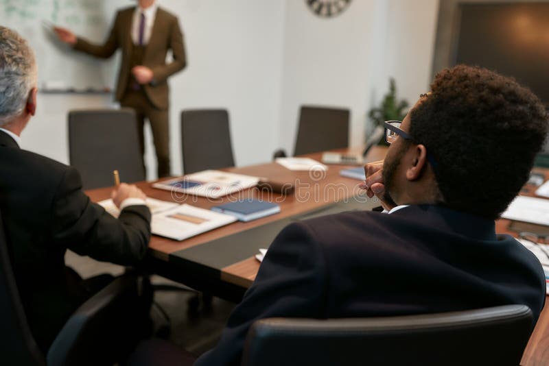 Foto recortada de jovem empresário em vestimenta clássica apontando para o quadro branco e explicando algo para o seu imagem de stock