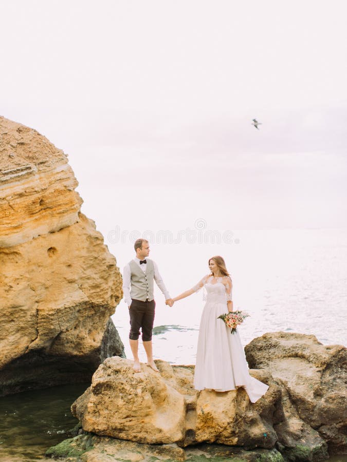 Foto verticale della mano della tenuta delle coppie della persona appena sposata sulla scogliera ai precedenti del mare fotografia stock Foto verticale della mano della tenuta delle coppie della persona appena sposata sulla scogliera ai precedenti del mare fotografia stock
