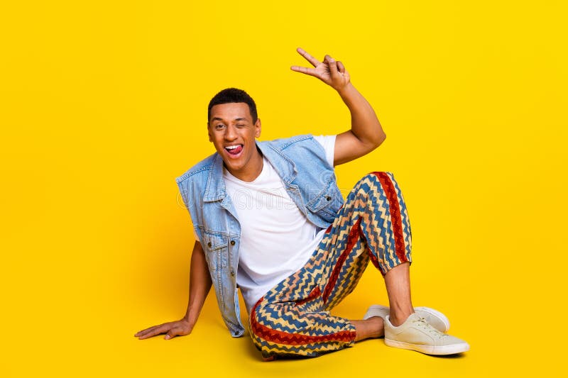 Full length photo of cheerful guy dressed jeans waistcoat sitting on floor winkin eye show v-sign isolated on yellow color background. Full length photo of cheerful guy dressed jeans waistcoat sitting on floor winkin eye show v-sign isolated on yellow color background.