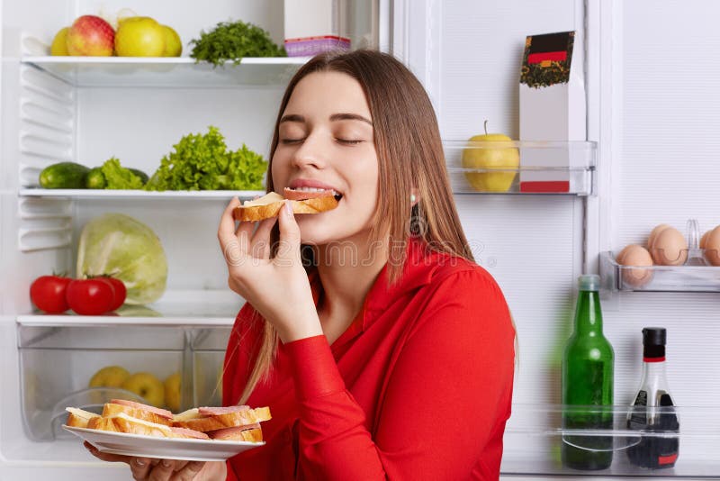 Fotoet av den hungriga unga gulliga kvinnan äter med den smakliga korvsmörgåsen för aptit, kommer efter arbete, står nära det öpp royaltyfria bilder Fotoet av den hungriga unga gulliga kvinnan äter med den smakliga korvsmörgåsen för aptit, kommer efter arbete, står nära det öpp royaltyfria bilder