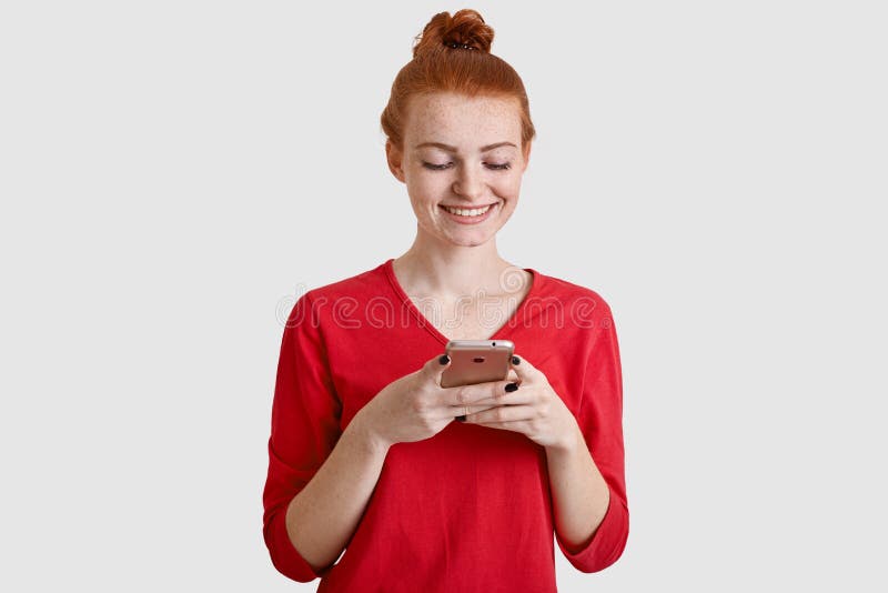 Fotoet av den tillfredsställda röda haired kvinnan med fräknig hud, rymmer den moderna mobiltelefonen, överför textmeddelandet, a royaltyfri foto Fotoet av den tillfredsställda röda haired kvinnan med fräknig hud, rymmer den moderna mobiltelefonen, överför textmeddelandet, a royaltyfri foto