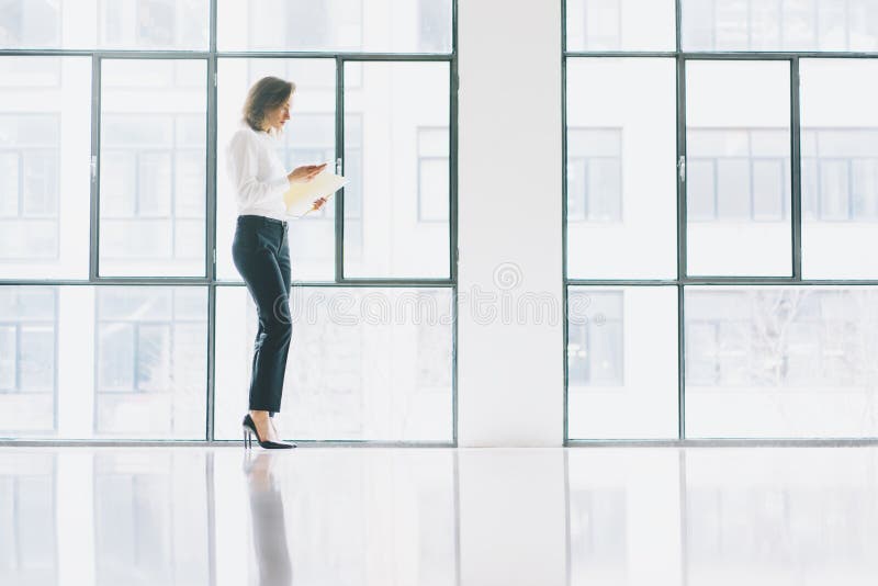 FotoGeschäftsfrau, die modernen Anzug trägt, Handy schaut und Papiere in den Händen hält Dachbodenbüro des offenen Raumes lizenzfreie stockfotos FotoGeschäftsfrau, die modernen Anzug trägt, Handy schaut und Papiere in den Händen hält Dachbodenbüro des offenen Raumes lizenzfreie stockfotos