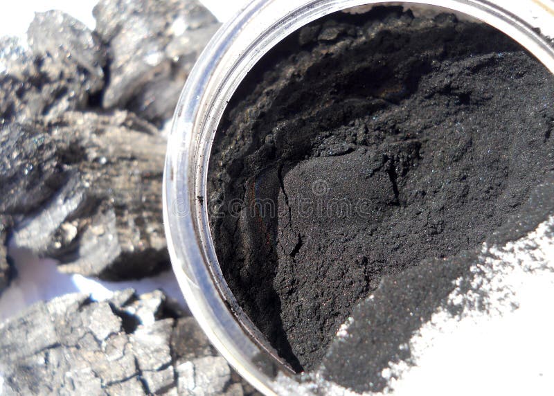 Macro photograph of jar of black activated charcoal powder. Background shows pieces of hardwood charcoal. Macro photograph of jar of black activated charcoal powder. Background shows pieces of hardwood charcoal.