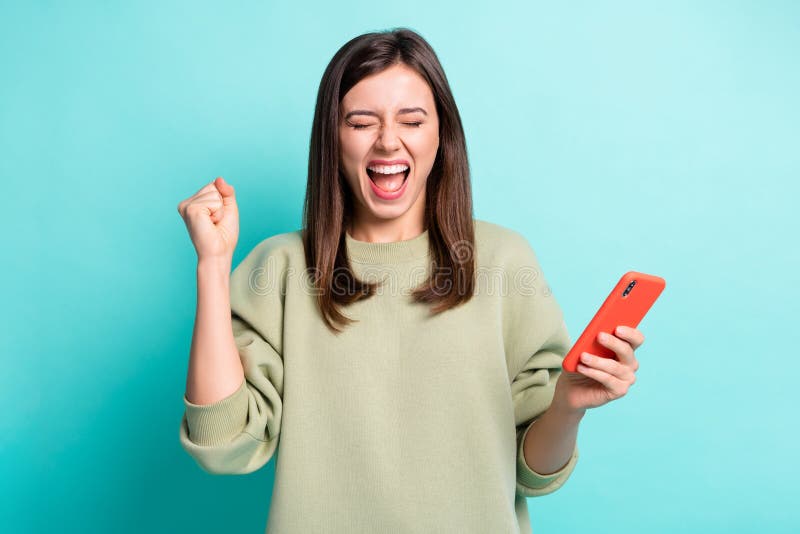 Photo portrait of crazy screaming girl raising fist up holding phone in one hand isolated on vivid cyan colored background. Photo portrait of crazy screaming girl raising fist up holding phone in one hand isolated on vivid cyan colored background.