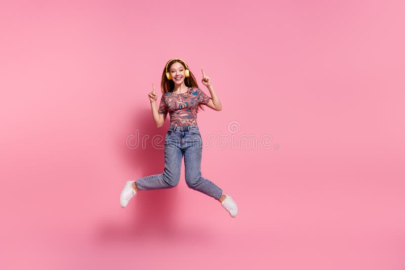 Full length photo of lovely teen lady jump headphones point up empty space dressed stylish print clothes isolated on pink color background. Full length photo of lovely teen lady jump headphones point up empty space dressed stylish print clothes isolated on pink color background.