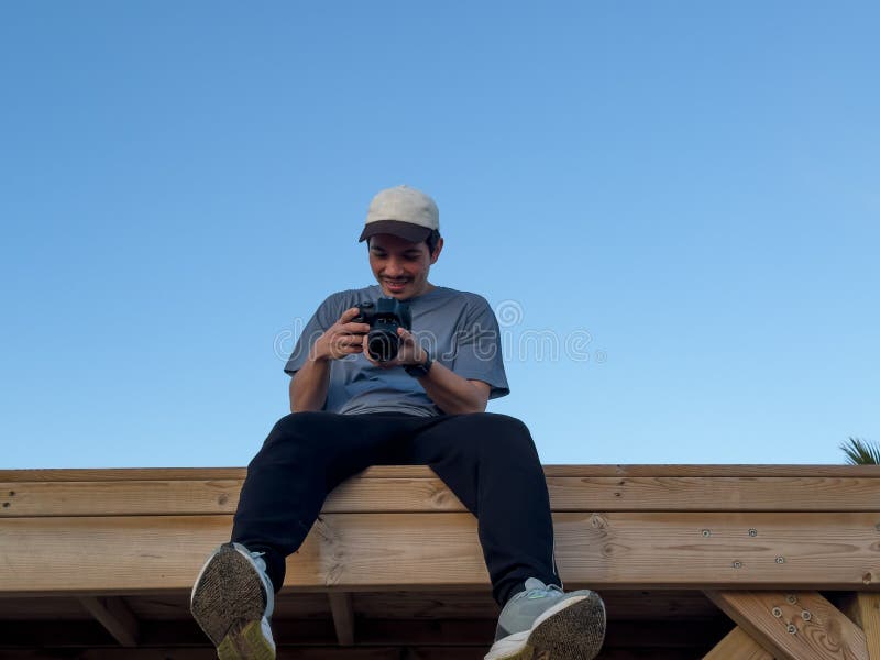 Fotógrafo sonriente sentado en una estructura de madera con un cielo azul al fondo mirando las fotos que ha tomado fotos de archivo libres de regalías